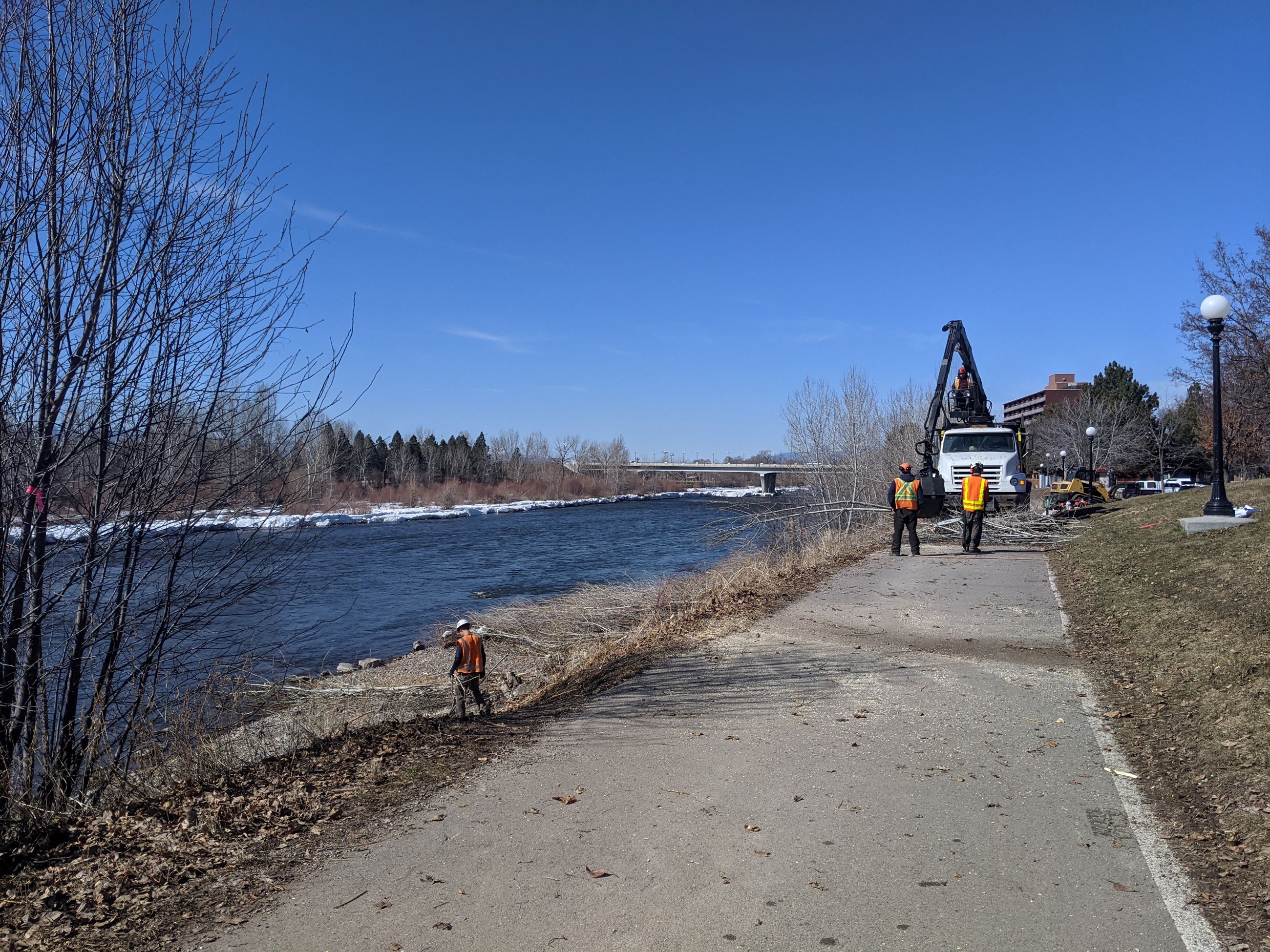 Large machine removing cut branches from the levee with workers in orange vests standing nearby.